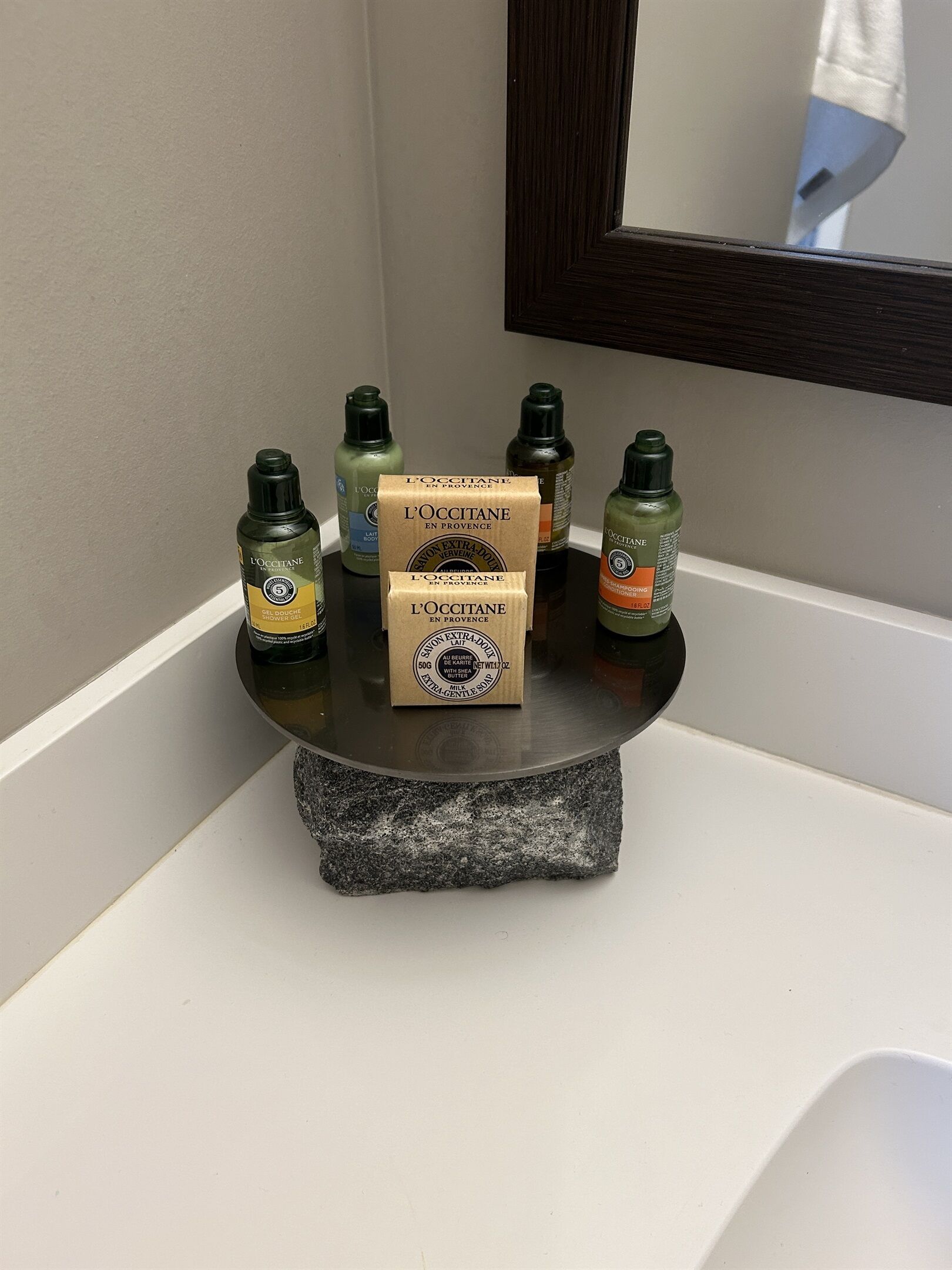 Een badkamer wastafel met een stenen aanrechtblad, een spiegel erboven en verschillende flesjes L'OCCITANE douchegel.
