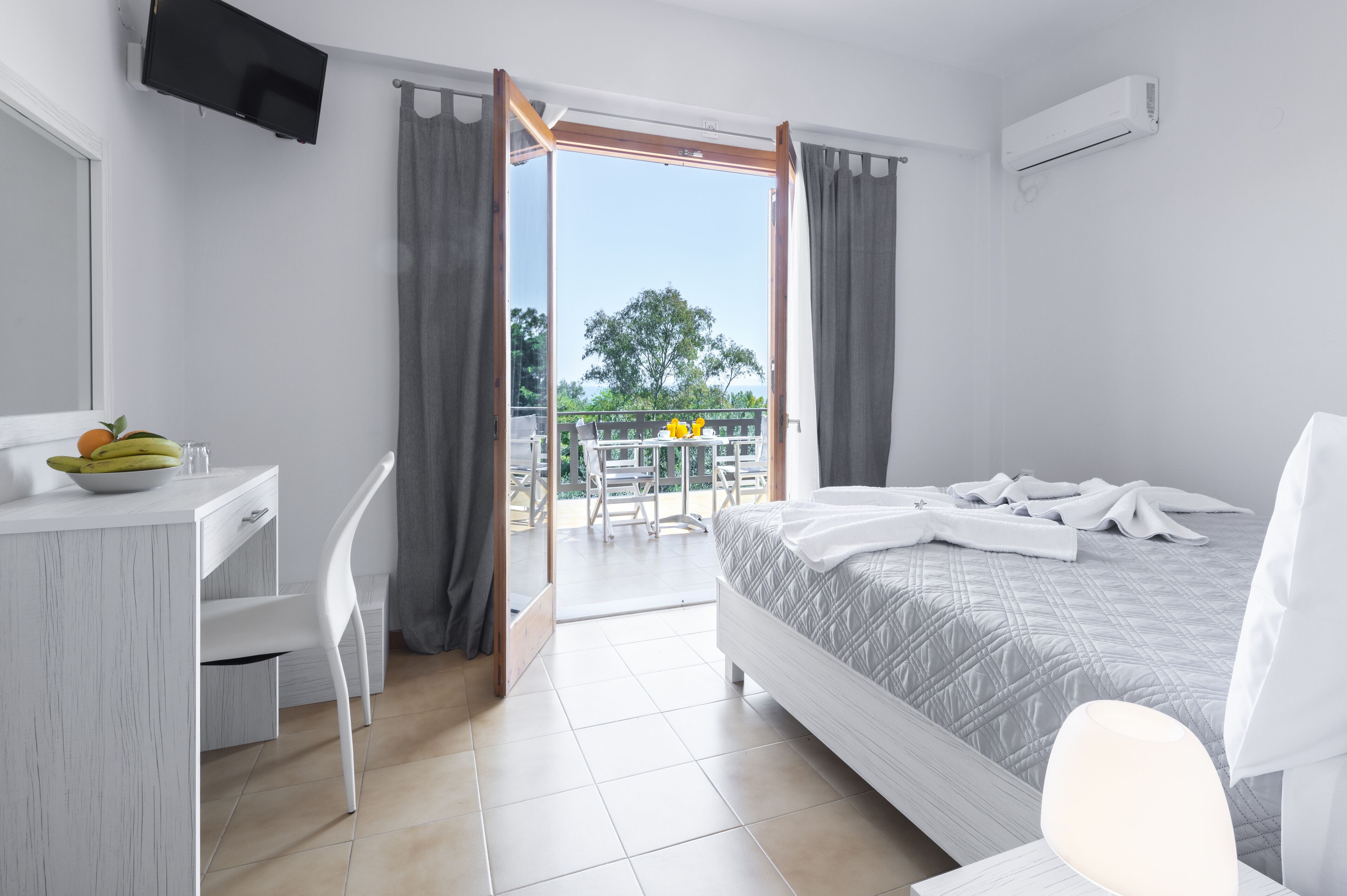 A hotel room with a bed, a desk with a bowl of fruit, a chair, and a balcony with a table and chairs.