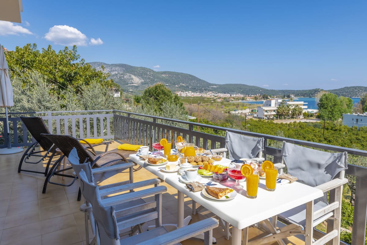 A terrace with a table set for breakfast, overlooking a scenic landscape with hills and a body of water.