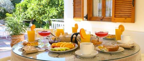 A breakfast table with various fruits, pastries, and beverages, set on a glass table with lace tablecloth, overlooking a garden with trees and a house.