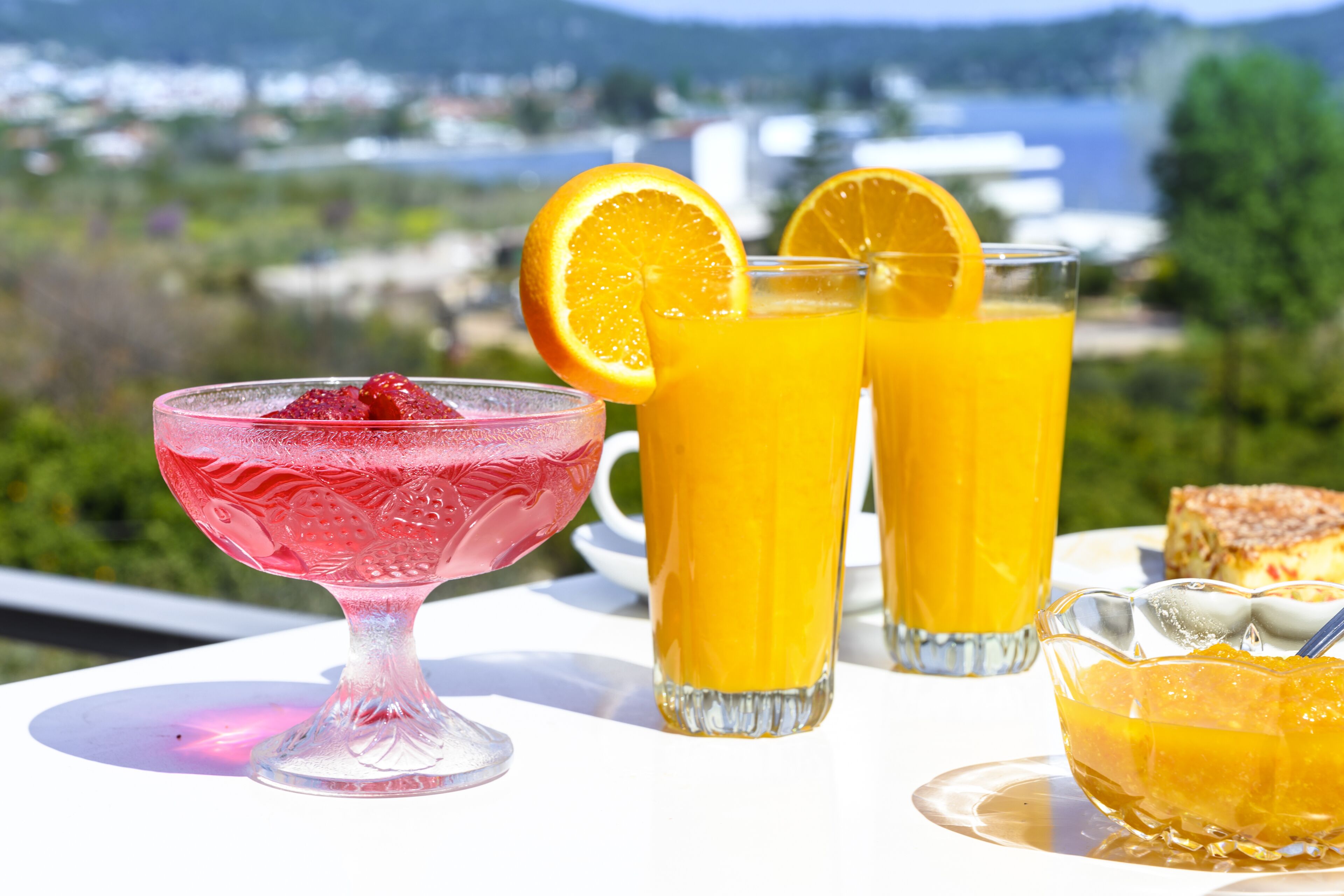 A table with a pink drink, two tall glasses of orange juice, and a bowl of fruit on a terrace with a scenic view.