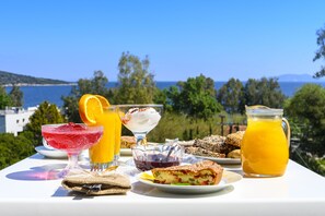 Una mesa con comida y bebidas, una jarra de vidrio con jugo de naranja, un tazón de bayas y un vaso de bebida roja, frente a una vista panorámica del mar y las colinas.