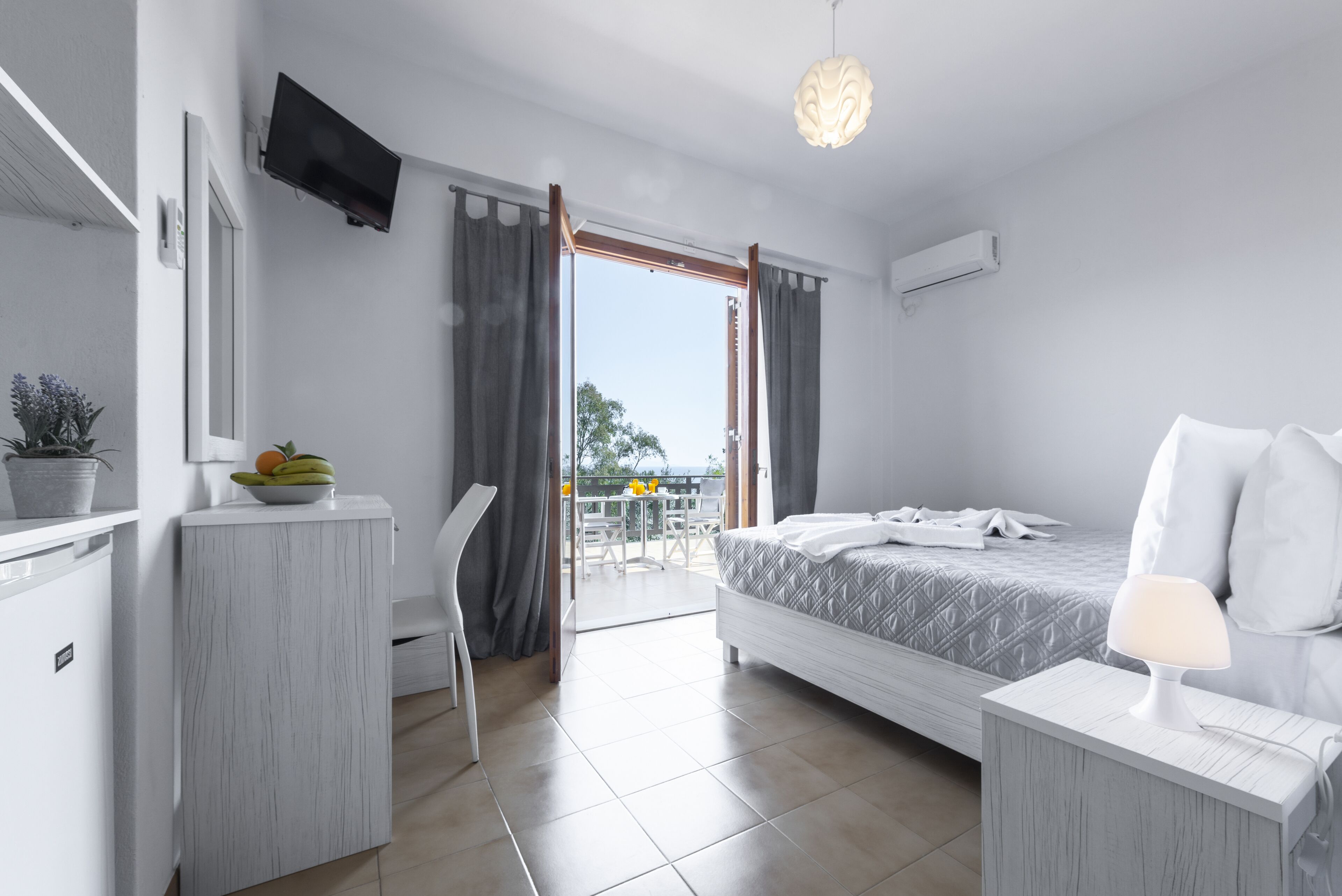 Una camera d'albergo moderna con un letto, un comodino e una scrivania con una sedia. Presenta un balcone con tavolo e sedie, e una televisione montata a parete.