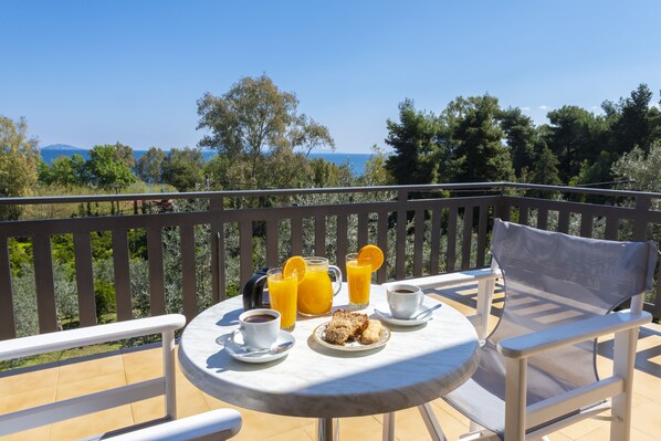 Una terraza con una mesa preparada para el desayuno, que incluye café, jugo de naranja y pastelería, con vistas a un paisaje pintoresco.