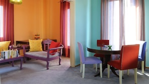 A room with a purple sofa, a small table with books, and a dining table with chairs. - Chroma Design Hotel and Suites (Nafplio)