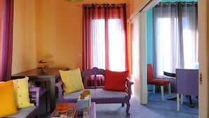 A cozy living room with a sofa, a dining table, and a chair. There is a window with curtains, a lamp, and a view of the outside. - Chroma Design Hotel and Suites (Nafplio)