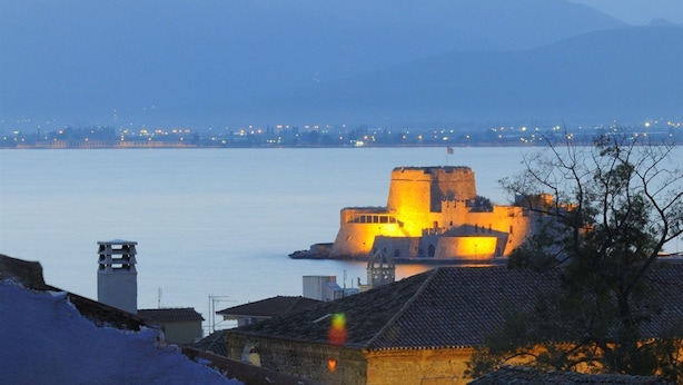 A table with a lantern, a drink, and a glass of water, overlooking a historic building by the water. - Chroma Design Hotel and Suites (Nafplio)