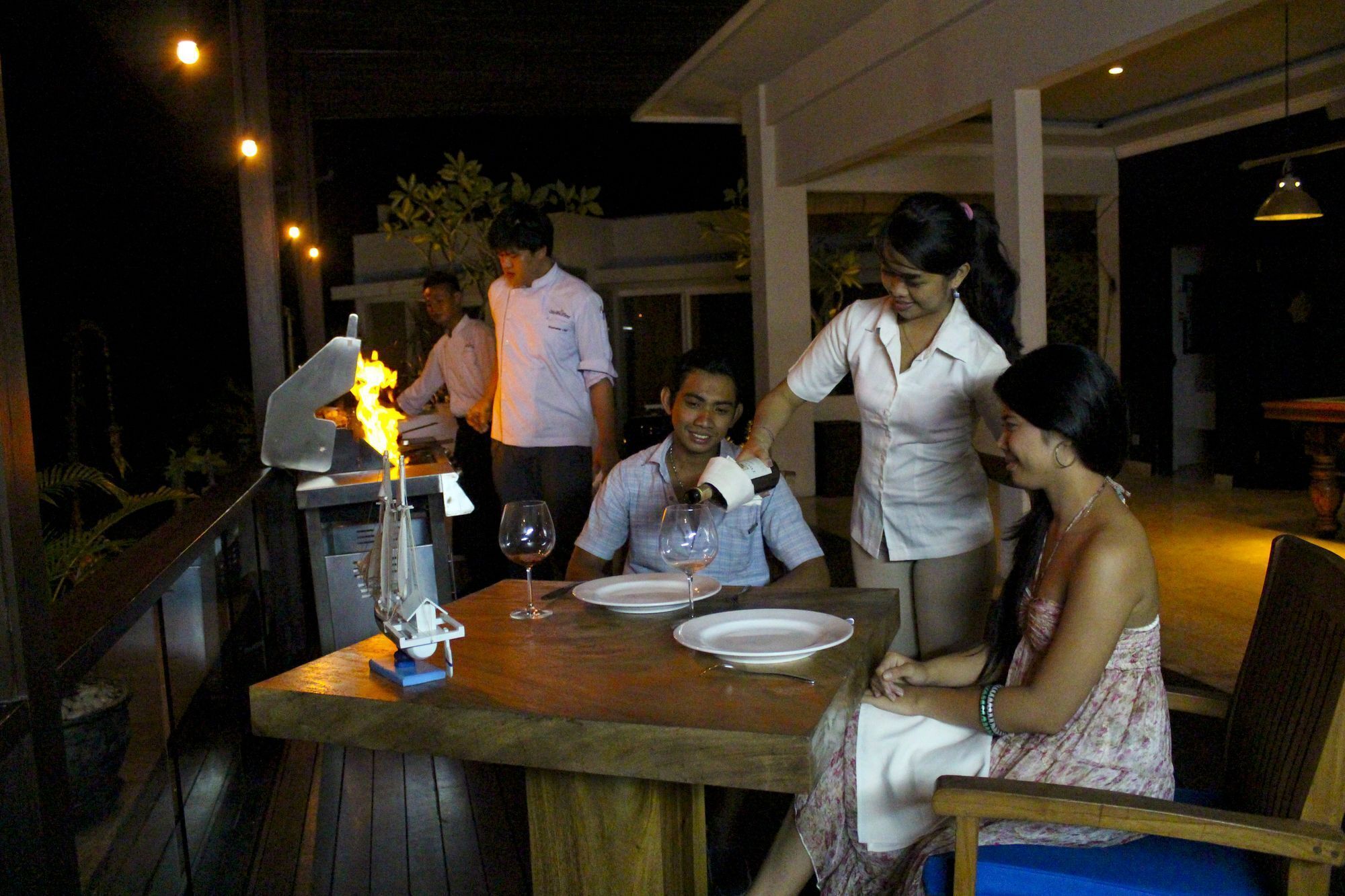 An outdoor dining area with a wooden table, a grill with flames, and a wine glass.