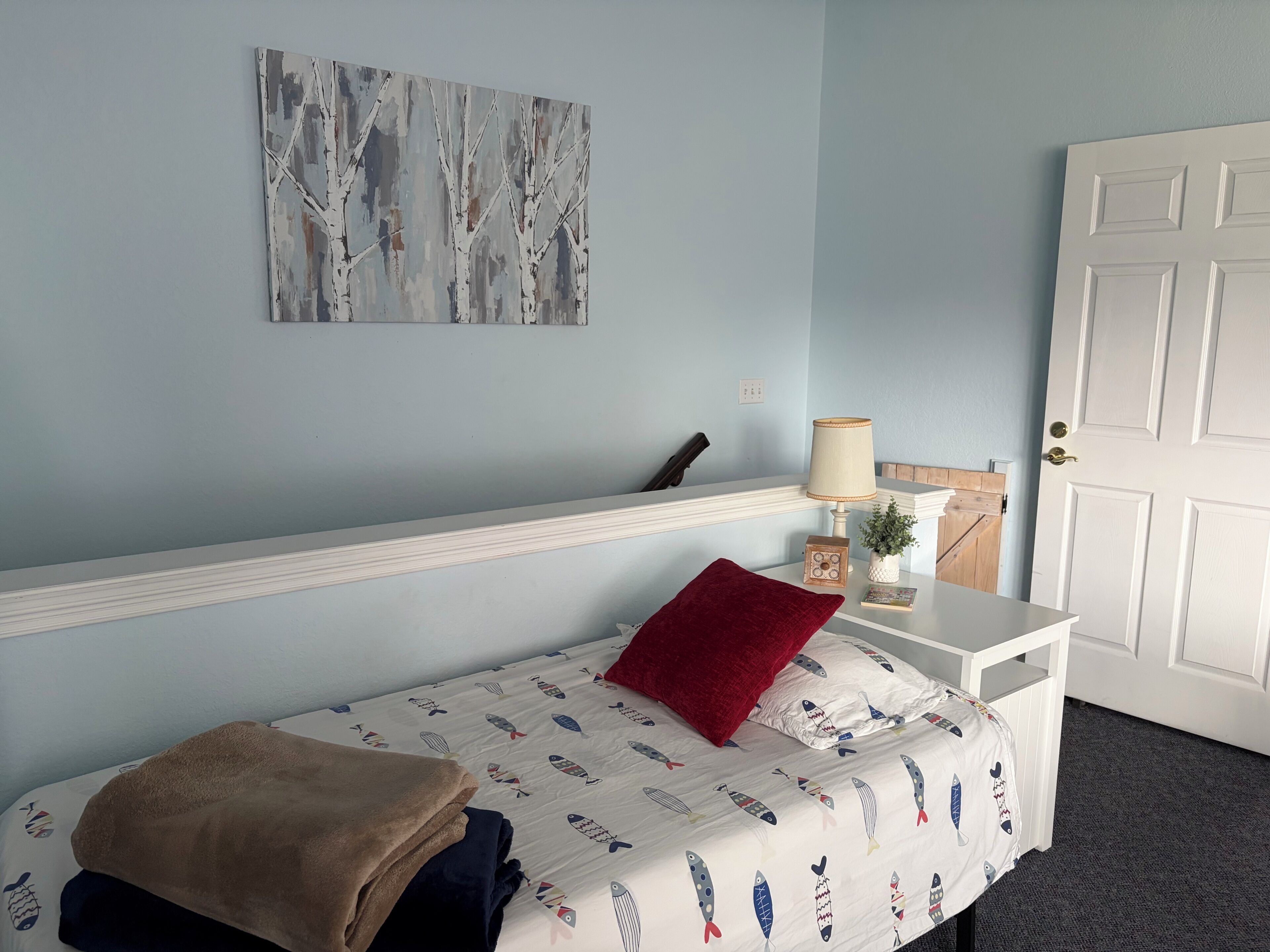 A bedroom with a bed, a red pillow, a lamp, a small plant, and a painting on the wall.