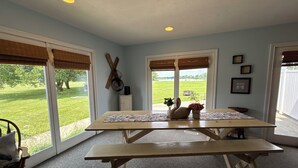 Ein Zimmer mit einem großen Tisch, zwei Jalousiewindosen, einer Tür und Blick auf ein grünes Feld.