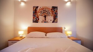 Una cama bien hecha con sábanas blancas, dos lámparas de noche y una obra de arte en la pared con un árbol.