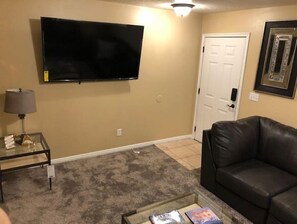 Ein Wohnzimmer mit einem an der Wand befestigten Flachbildfernseher, einem Glastisch mit Büchern, einem schwarzen Ledersofa und einer Tür mit weißem Rahmen.