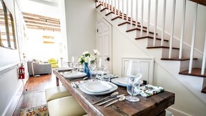 Un coin repas avec une table en bois dressée pour quatre personnes, des chaises blanches et un escalier en arrière-plan.
