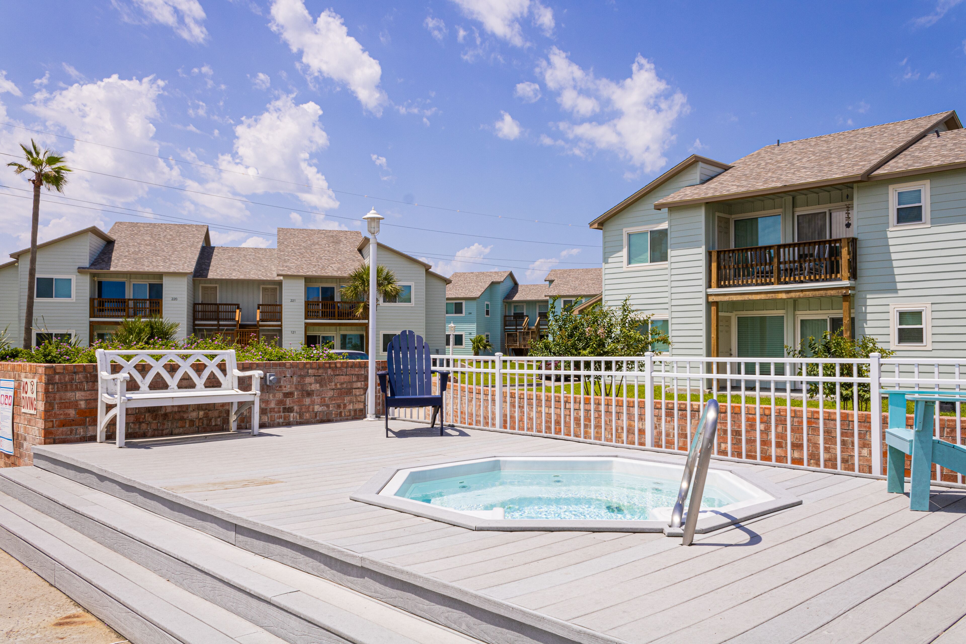 A pool area with a hot tub, wooden deck, and a white fence.