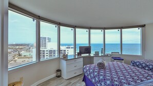Un dormitorio con un amplio ventanal, una cama con colcha estampada, un escritorio con computadora y vistas al mar y al paisaje urbano.