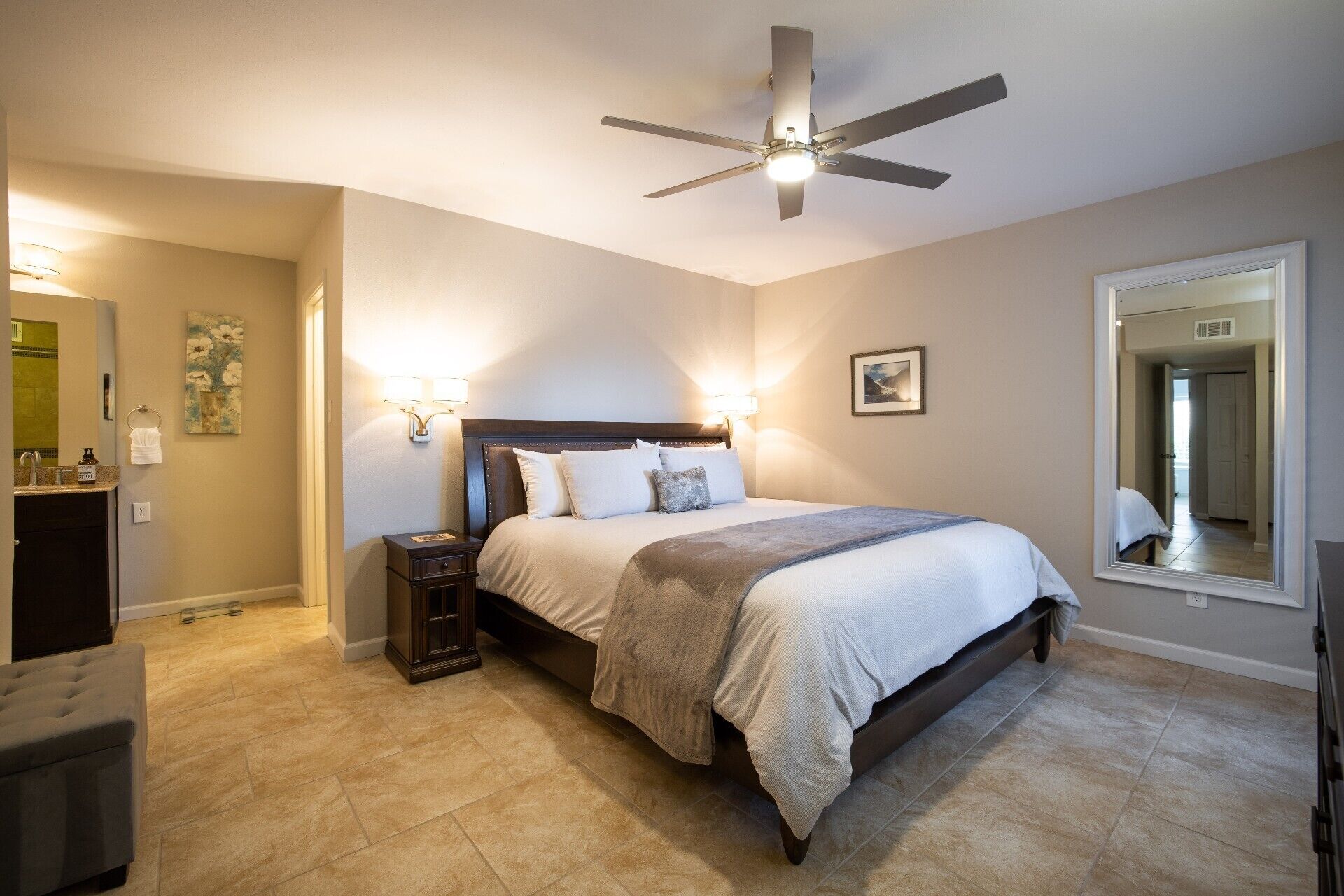 A bedroom with a bed, nightstands, a ceiling fan, a large mirror, and a bathroom with a sink and towel rack.