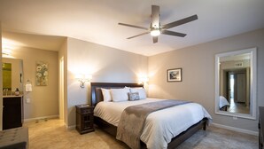 Une chambre à coucher comprenant un lit, des tables de chevet, un ventilateur de plafond, un grand miroir et une salle de bain avec un lavabo et un porte-serviettes.