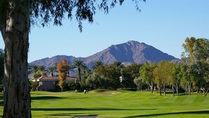 Un parcours de golf avec une montagne en arrière-plan, un grand arbre au premier plan et un bâtiment avec un toit rouge.