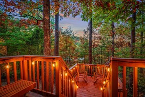 Une terrasse en bois, illuminée de guirlandes lumineuses, entourée d’arbres et offrant une vue sur une forêt.