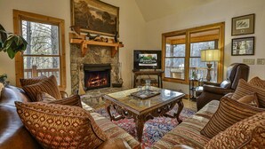 A cozy living room with a stone fireplace, leather furniture, and large windows. - Riverview Hideaway by Mountain Oasis | Dog-Friendly Riverview Cabin in Coosawattee (Ellijay)