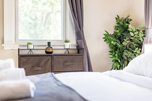 Een netjes opgemaakt bed met witte lakens, een nachtkastje met twee planten in potten, en een raam met gordijnen.