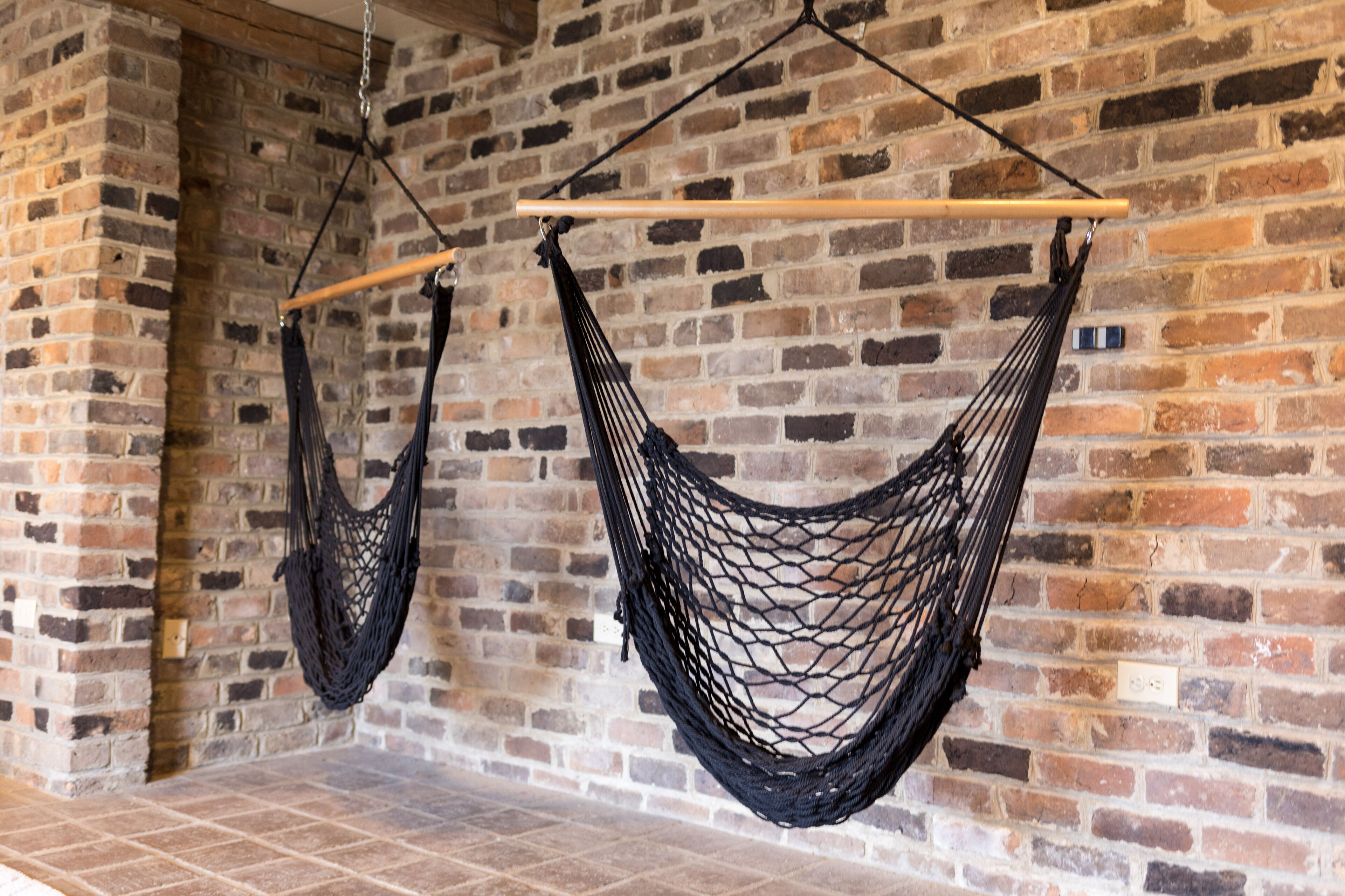Two black hammocks suspended from a wooden beam against a brick wall.