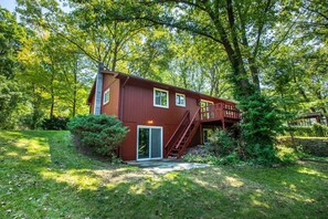 Une cabane en bois à deux étages, dotée d’une terrasse et entourée d’une végétation luxuriante et d’arbres denses.