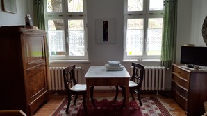 A room with a wooden cabinet, a table with chairs, a radiator, and a window with curtains. - Renovated farmhouse in the northern Uckermark, between lakes and forests (Brüssow)