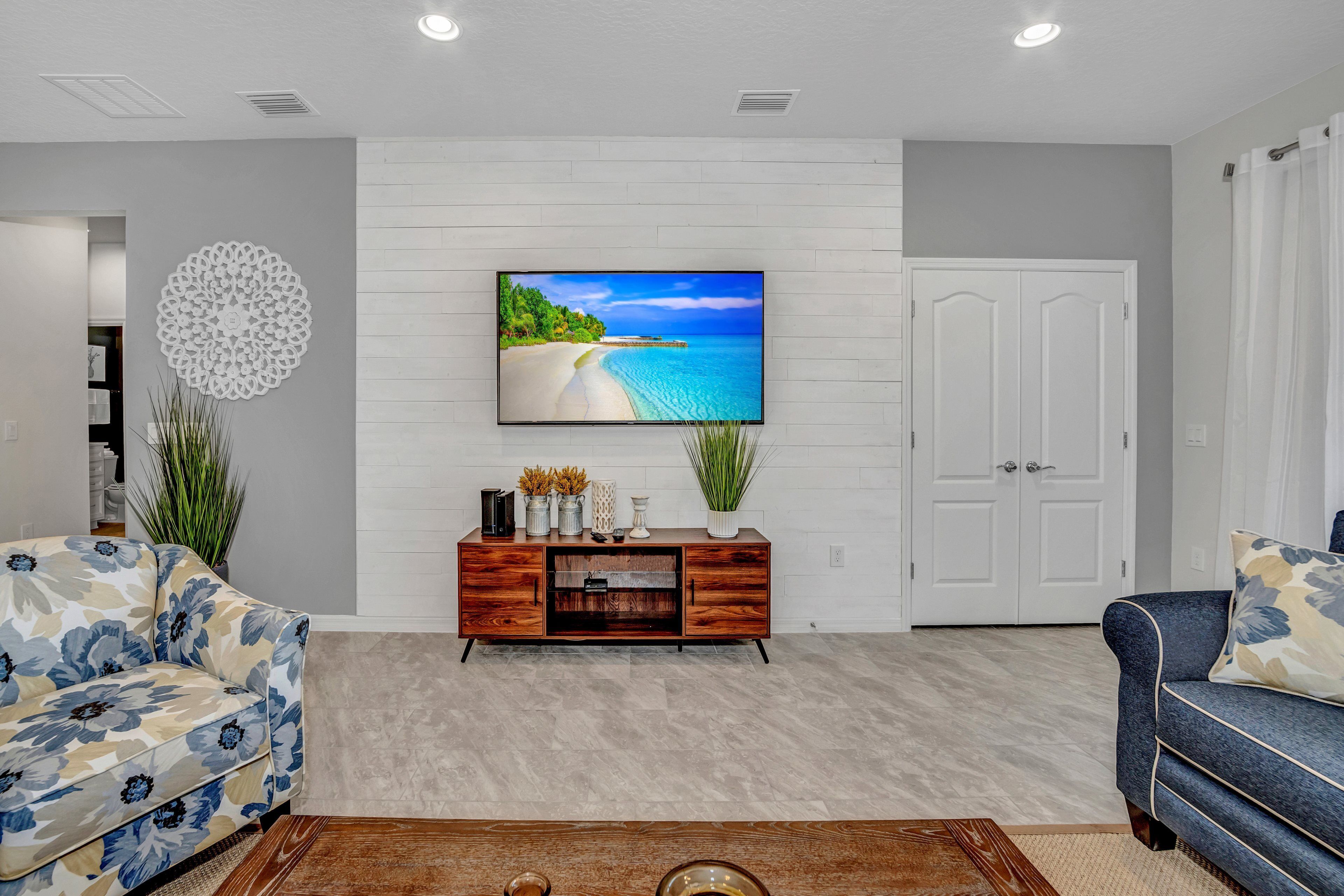 A modern living room with a large flat-screen TV mounted on a white brick wall, a wooden cabinet below, and two sofas with floral upholstery.