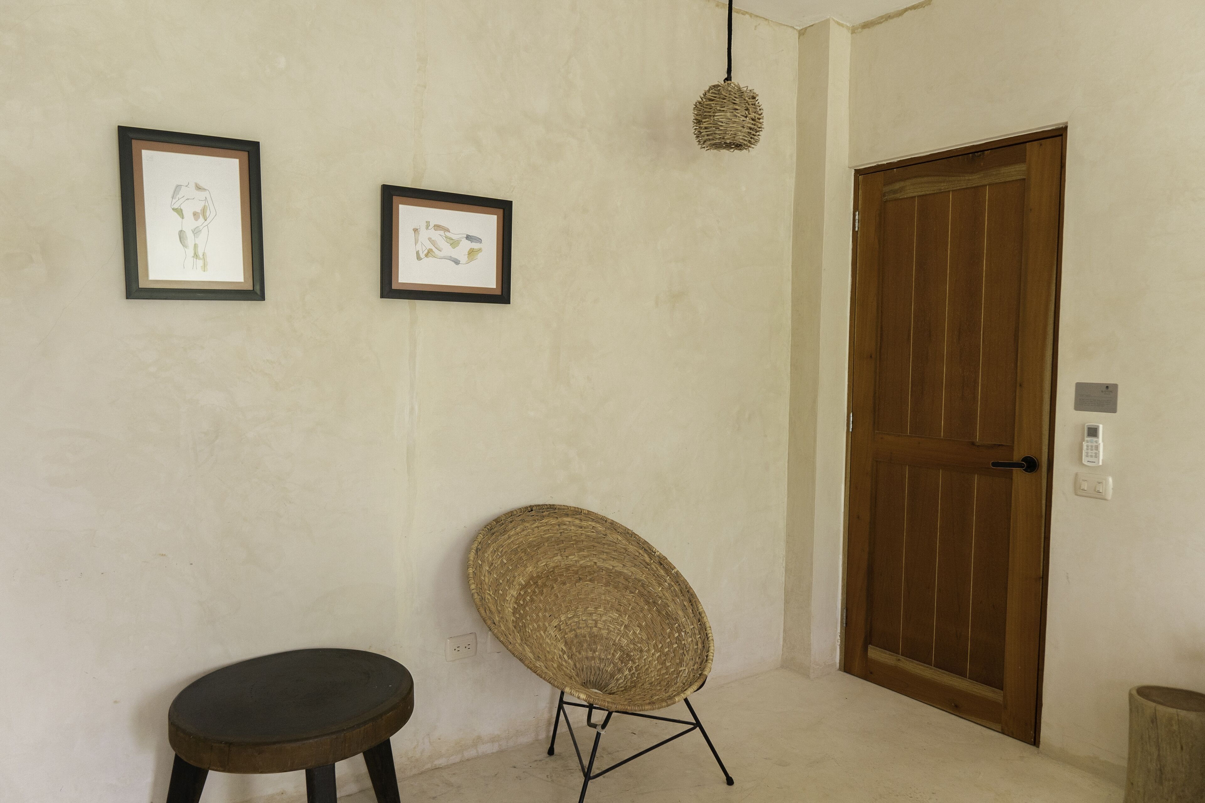 Ein Zimmer mit Holztür, zwei gerahmten Kunstwerken an der Wand, einem gewebten Stuhl und einem kleinen Hocker.