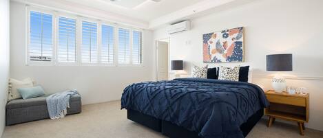 Quarto com cama grande, ventilador de teto, janela com persianas, ar condicionado fixado na parede e um quadro decorativo.