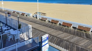 Blick auf einen Strand vom Balkon, mit Holzgeländern, weißen Liegestühlen und einem Schild, das einen Ort angibt.