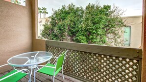 Un balcon avec une table en verre et deux chaises, donnant sur un buisson touffu et verdoyant ainsi que sur des immeubles.