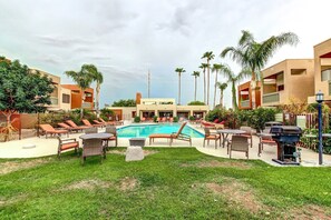 Zona de piscina con sillones de descanso y una parrilla, rodeada de palmeras y edificios modernos.