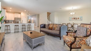 A spacious living area with a sofa, coffee table, and bar stools. A kitchen is visible in the background with a refrigerator and a dining area with chairs. - C9052sid- The Retreat at Champions Gate (Davenport)
