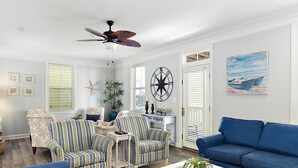 Un salon avec des fauteuils à rayures, un canapé bleu, un ventilateur de plafond, une horloge et un tableau sur le thème de la marine.