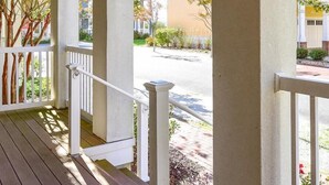Une véranda couverte avec des balustrades blanches et un plancher en bois, donnant sur une rue bordée d’immeubles.