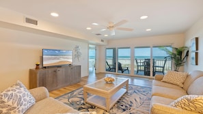 A living room with a TV, a ceiling fan, a balcony with a view of the beach, and a large window.