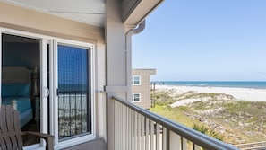 A balcony with a view of the beach, a bed with blue bedding, and a chair.