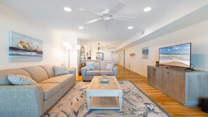 A spacious living area with a sofa, coffee table, and TV, a kitchen in the background, and a ceiling fan.