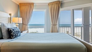 A hotel room with a large bed, a nightstand, a lamp, and a view of the beach.