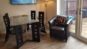 A modern living room with a glass dining table, black chairs, a black leather sofa with a patterned throw, a floor lamp, and a TV displaying 'TOSHIBA'. - Caspian House (4 Bedrooms) - Serviced Accommodation (Grimsby)