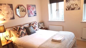 A bedroom with a large bed, a bedside table with a lamp, a mirror, and wall art including a peacock feather and floral paintings. - Caspian House (4 Bedrooms) - Serviced Accommodation (Grimsby)