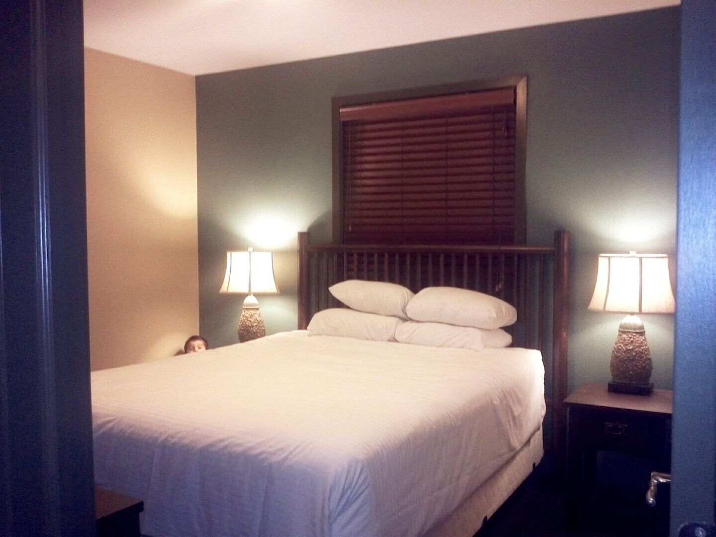 A hotel room with a large bed, two bedside tables with lamps, a window with blinds, and a wall-mounted headboard.