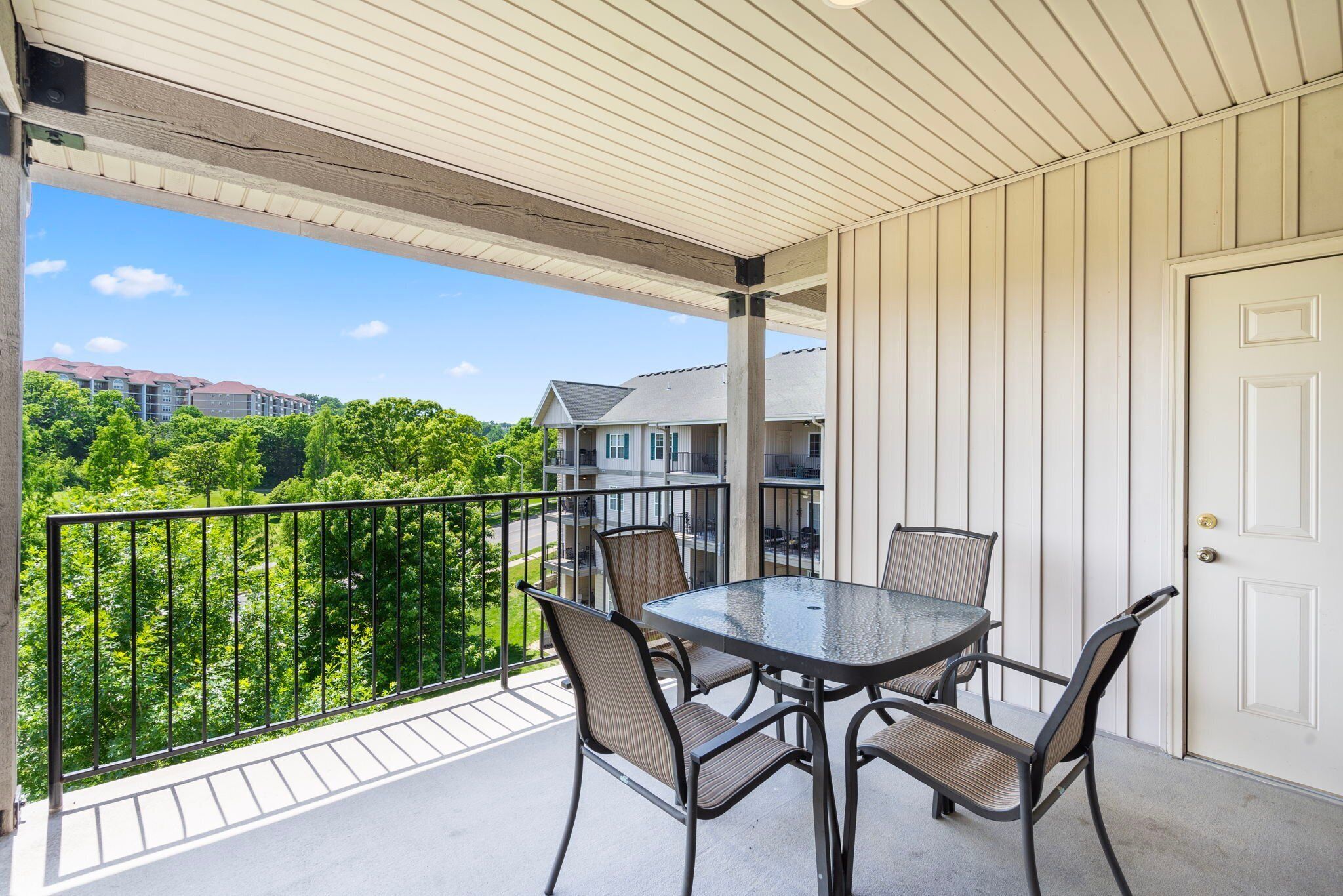 A covered balcony with a table and chairs, a white door, and a view of a residential area with trees and buildings.