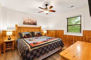 A bedroom with a wooden headboard, a bed with a quilt, two bedside lamps, a wooden nightstand, a wooden table, a ceiling fan, a window with blinds, and a wall-mounted picture. - Next to Rocky Top Sports! 5 King bedrooms 4 Bath Family-Friendly Sleeps 18 (Gatlinburg)