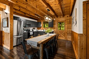 A wooden interior with a kitchen area featuring a stainless steel refrigerator, a microwave, and a stove. There is a dining table with chairs and a hanging light fixture above it. - Next to Rocky Top Sports! 5 King bedrooms 4 Bath Family-Friendly Sleeps 18 (Gatlinburg)