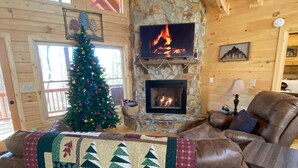 Interior acogedor de una cabaña con una chimenea de piedra, un televisor montado encima, un árbol de Navidad y un sofá de cuero con colcha.