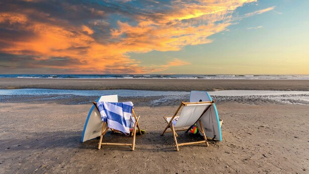 Zwei Strandstühle mit Surfbrettern an einem sandigen Strand, Blick auf das Meer und einen Sonnenuntergang.