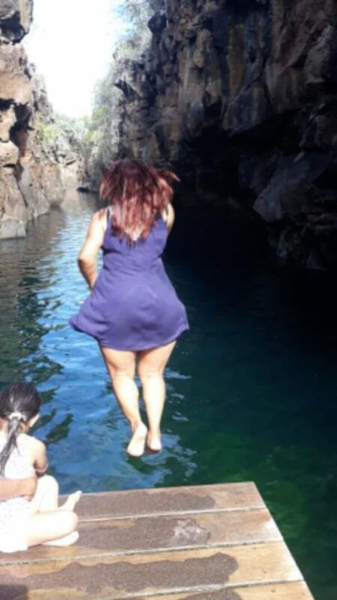 Een vrouw in een paarse jurk staat aan de rand van een houten steiger boven het water, met rotsachtige kliffen op de achtergrond.